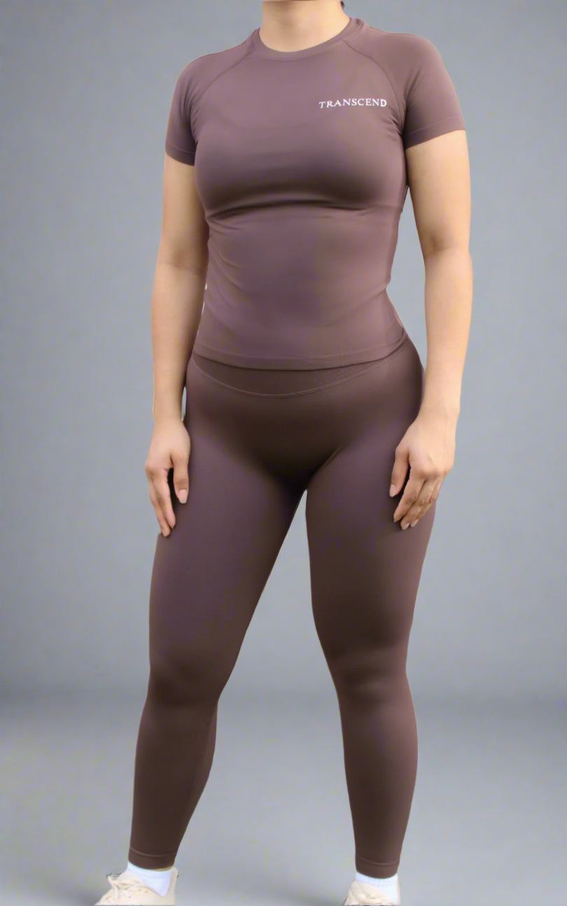 woman wearing brown athletic top with logo and brown legging standing in front of brick wall