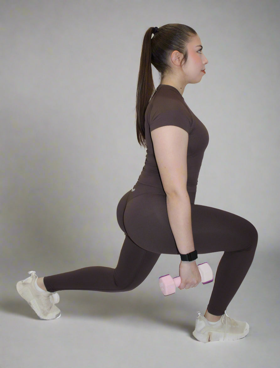 Woman in brown athletic wear performing a lunge with a pink dumbbell on a white background