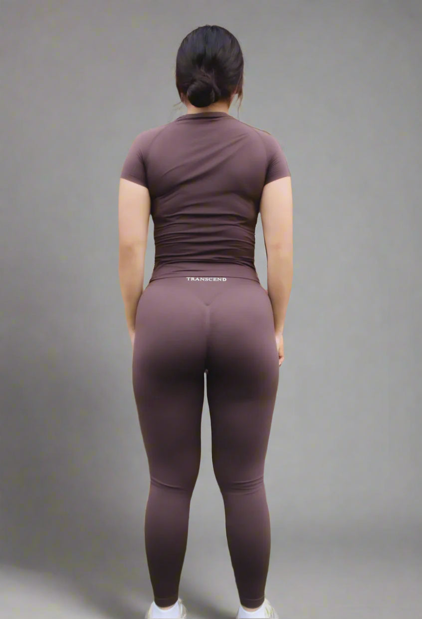 Person wearing brown athletic outfit with "TRANSCEND' branding against a brick wall.